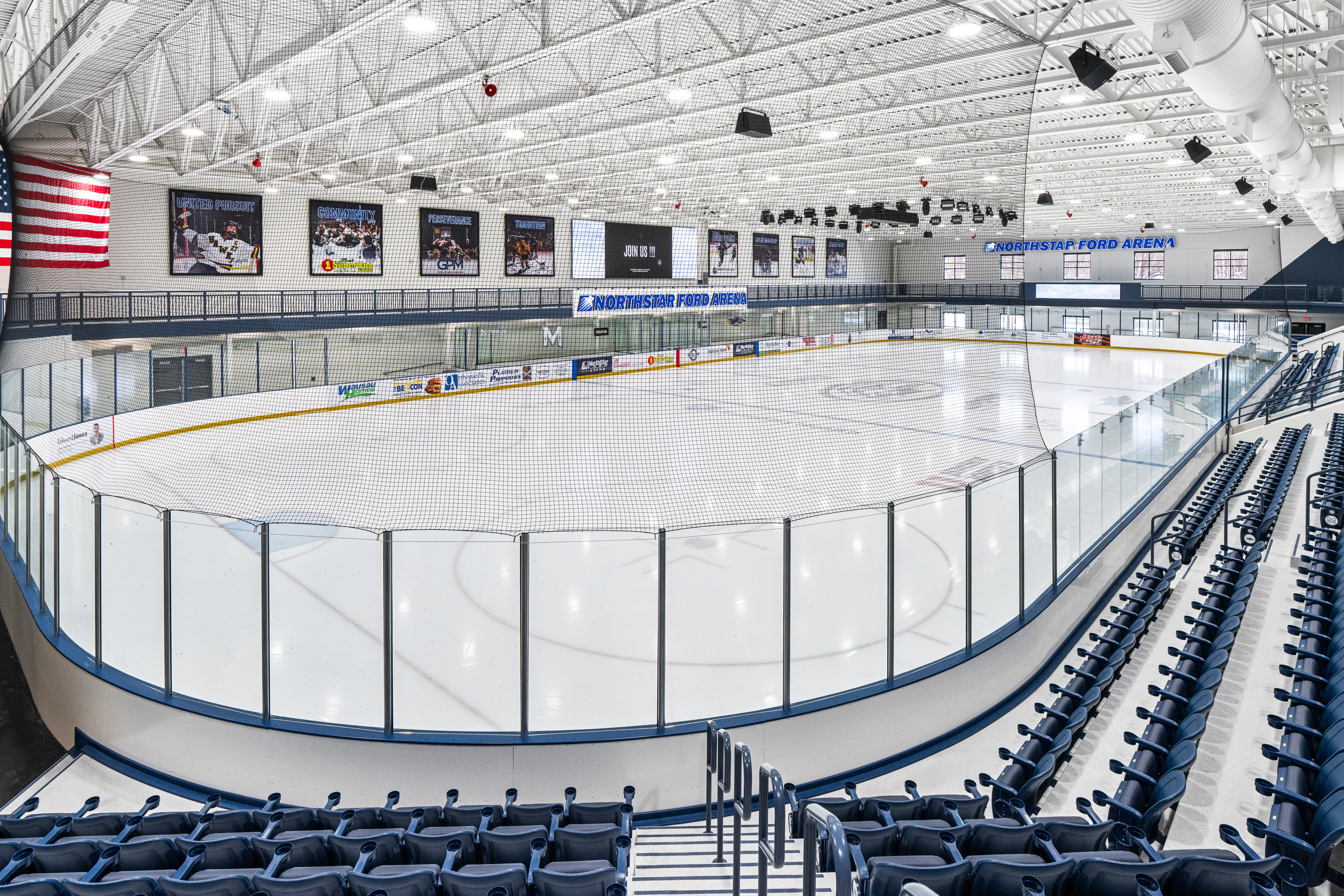 Empty indoor ice hockey rink with clear glass boards, blue spectator seats, overhead lights, an American flag on the left wall, and banners hanging above the ice at the end, reading "INDUSTRIAL FOAM ARENA.