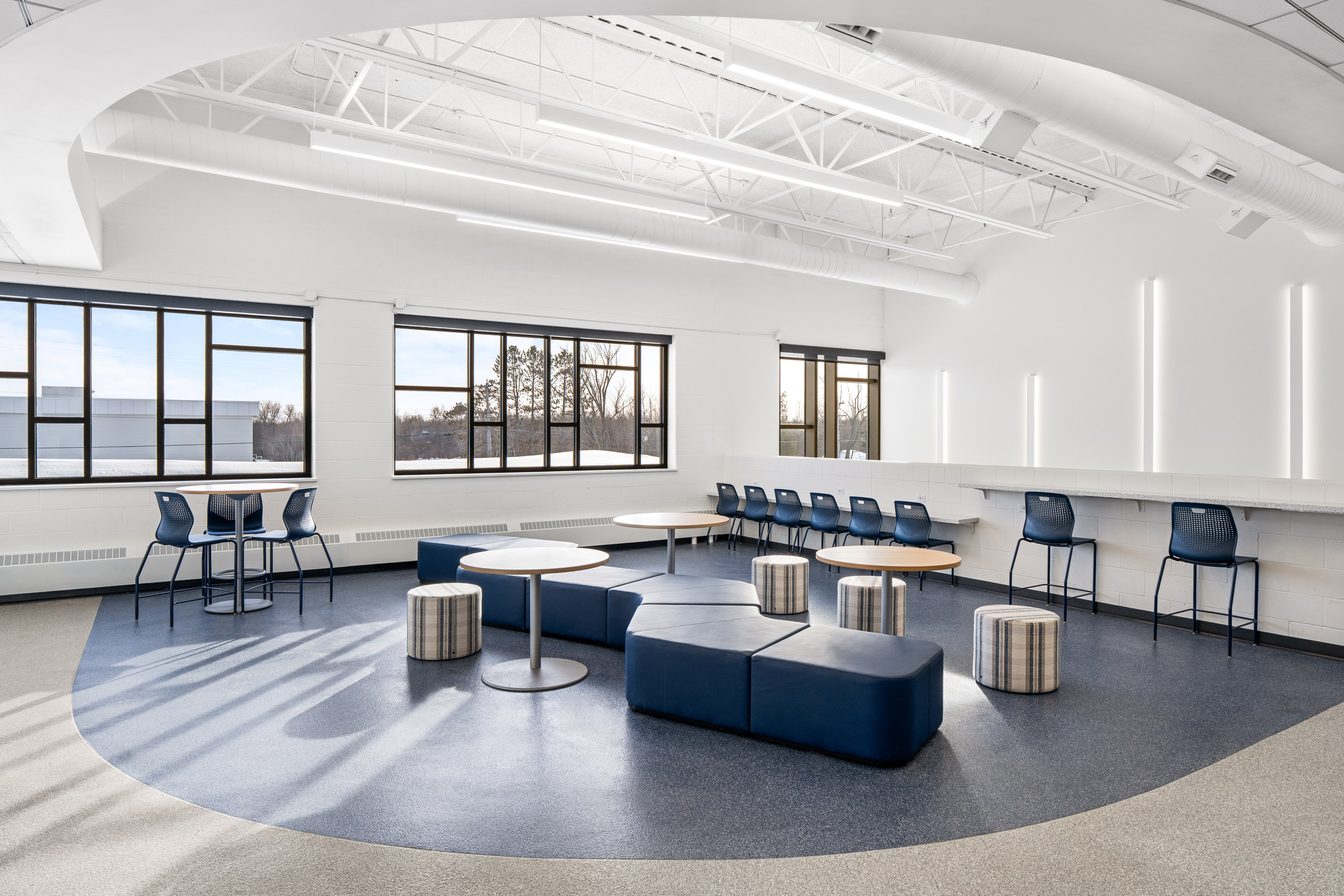 A modern, spacious room with large windows, blue and gray furniture, round tables, blue chairs, and high ceilings with exposed beams. Natural light fills the space, creating a bright and airy atmosphere.
