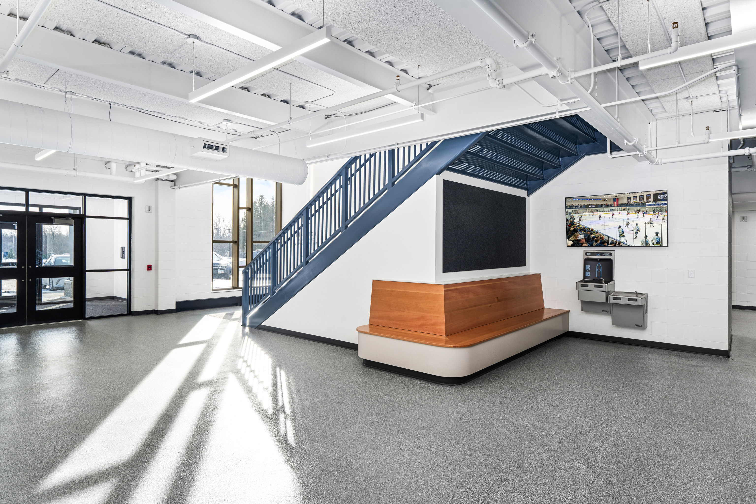 A bright, modern lobby with large windows, a blue staircase, a wooden bench, a wall-mounted TV displaying a hockey game, and two water fountains on a clean, gray floor.