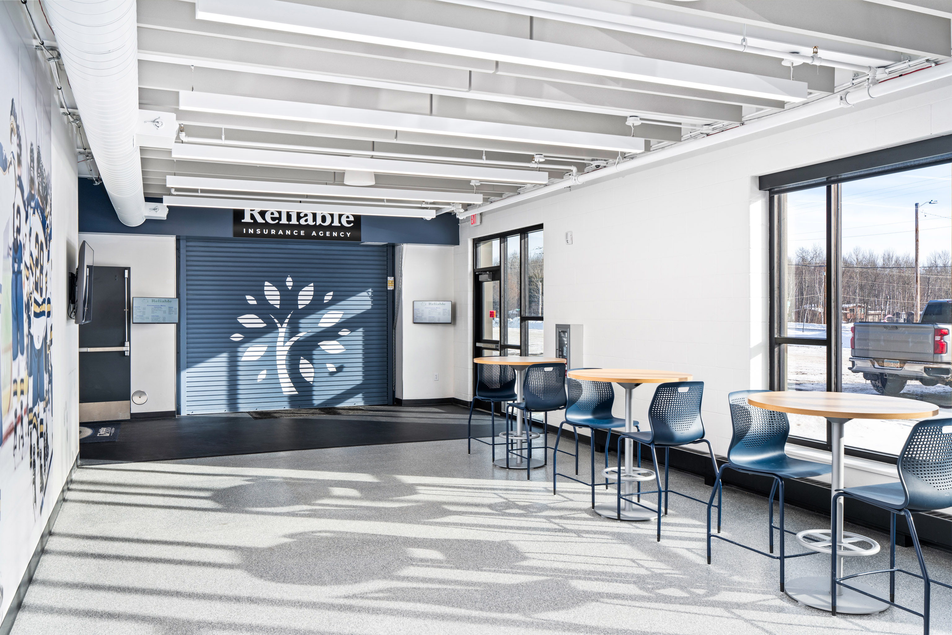 A bright, modern room with large windows, several round tables and blue chairs, and a wall featuring the "Reliable Insurance Agency" logo near a blue roll-up door. Snowy outdoors visible through the windows.