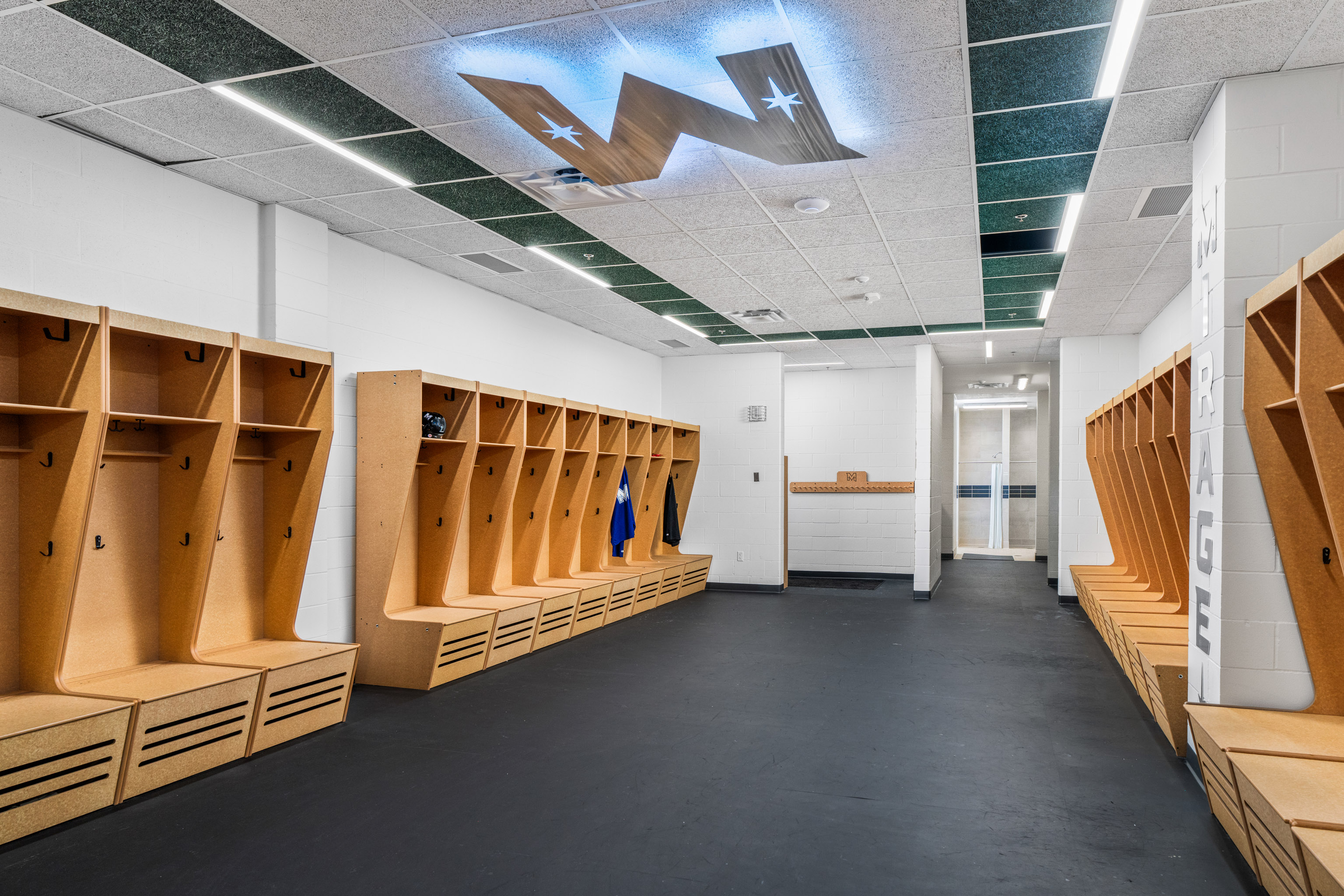 Modern locker room with wooden cubbies lining both sides, dark flooring, white walls, a counter at the far end, and a ceiling with a large illuminated “W” logo and star shapes.
