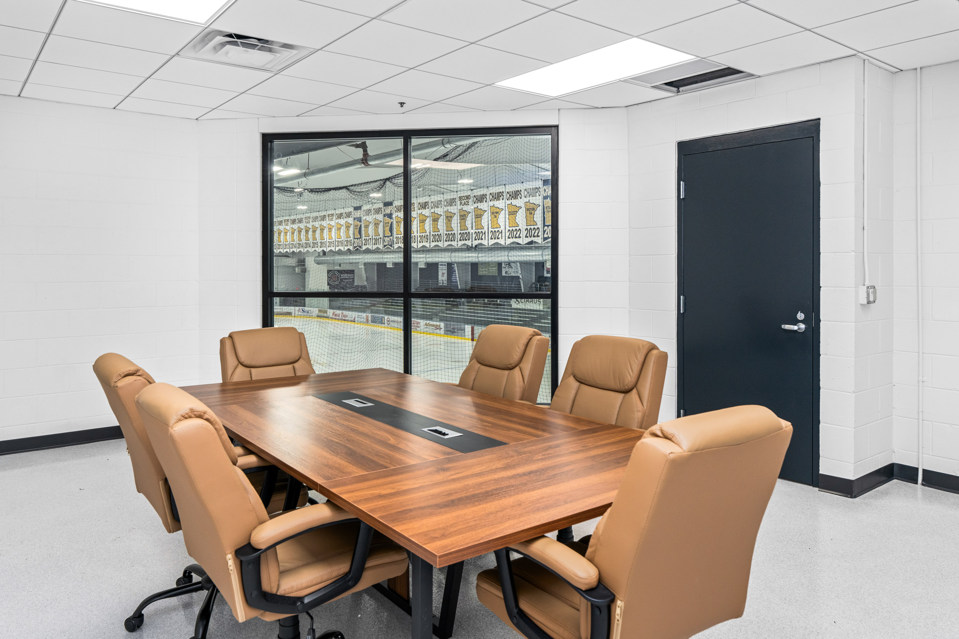 A modern conference room with a wooden table, six tan leather chairs, and a large window overlooking an ice rink with banners. The walls are white, and there is a closed dark blue door on the right.