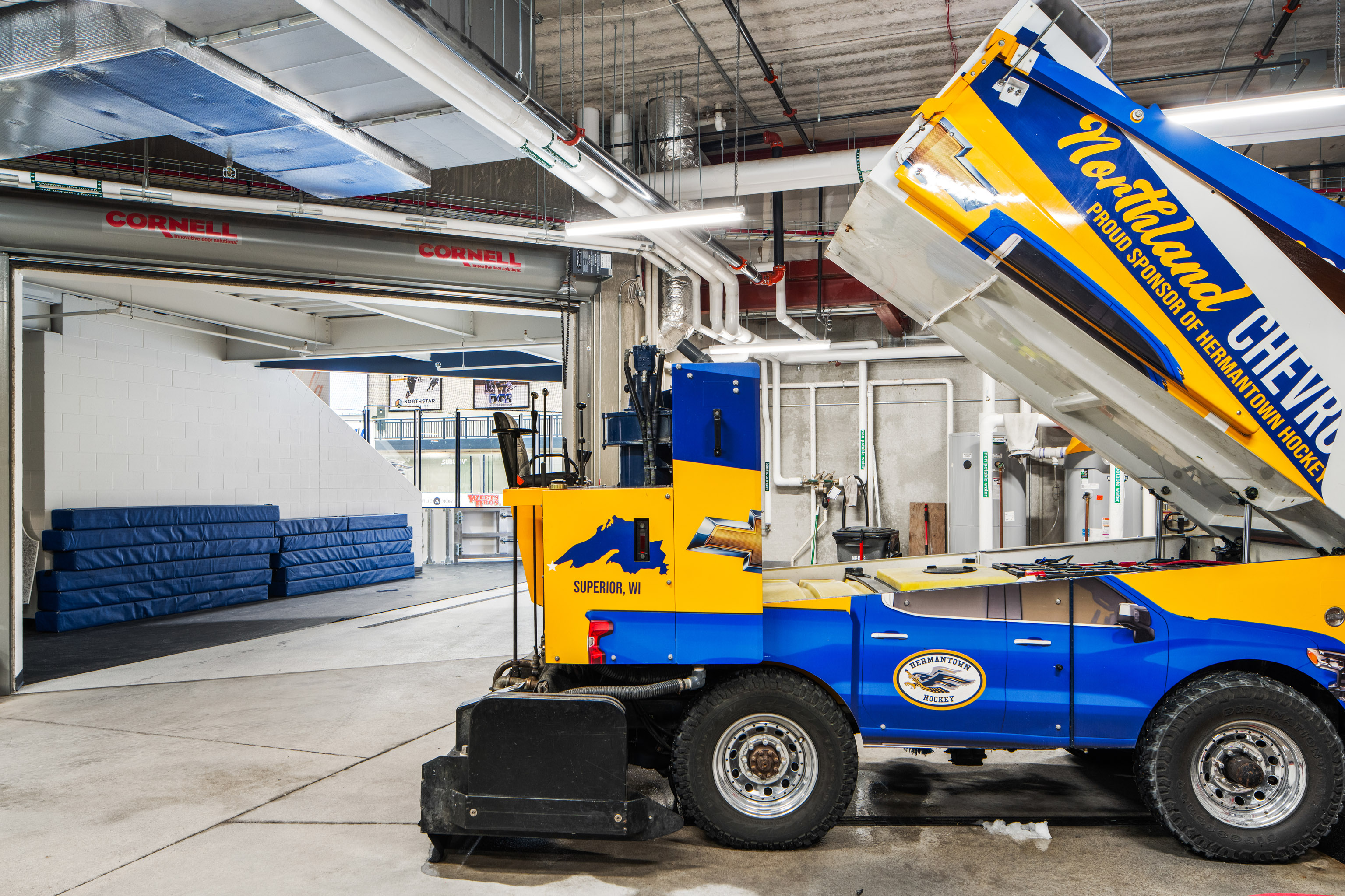 A blue and yellow ice resurfacing machine is parked indoors near stacked blue mats and an open garage door labeled “CORNELL.” The machine’s snow collection bin is raised. Pipes and lockers are visible in the background.