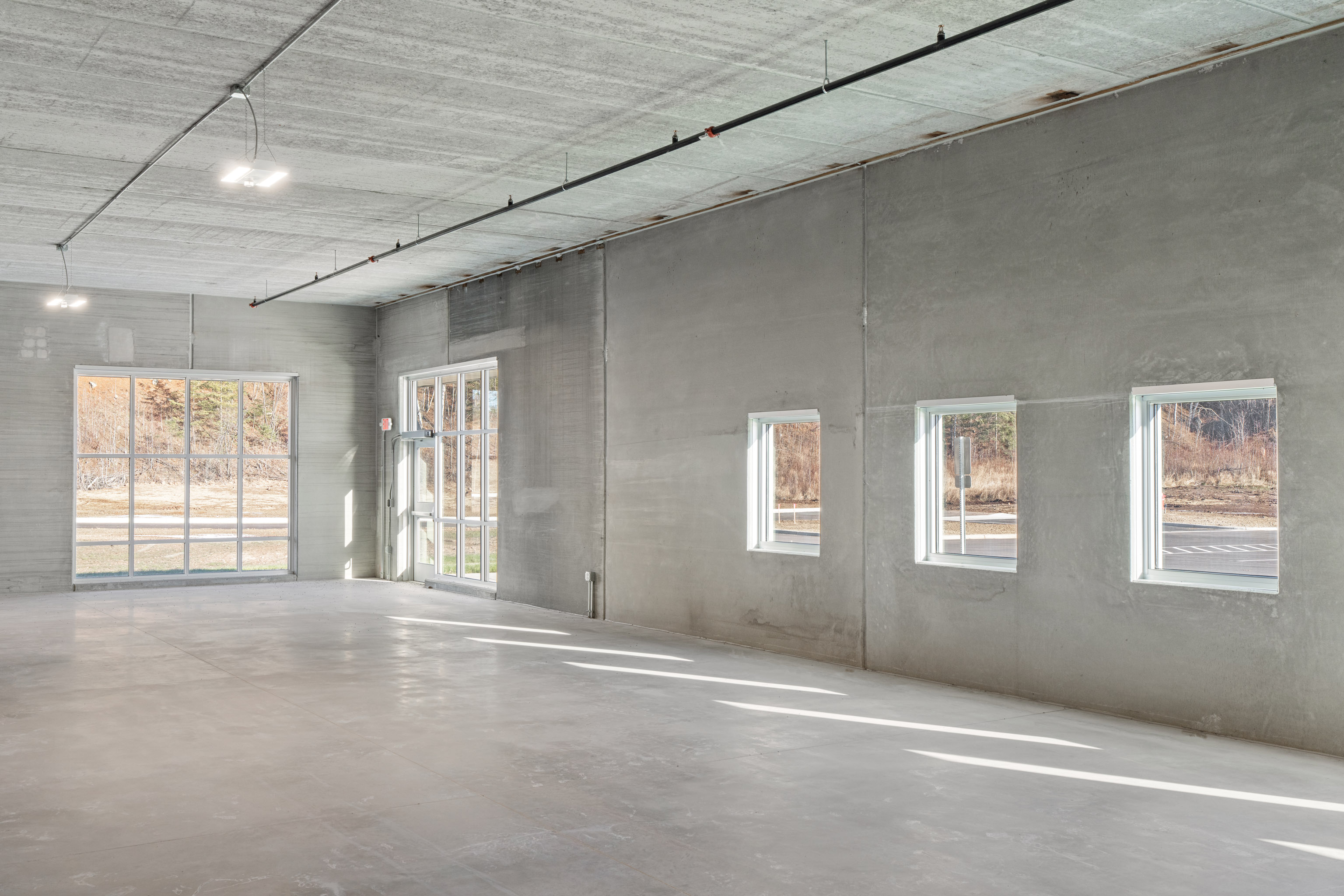 Empty industrial space with gray concrete floors and walls, large windows on the left, and three smaller windows on the right letting in natural light. Ceiling lights and exposed piping are visible.