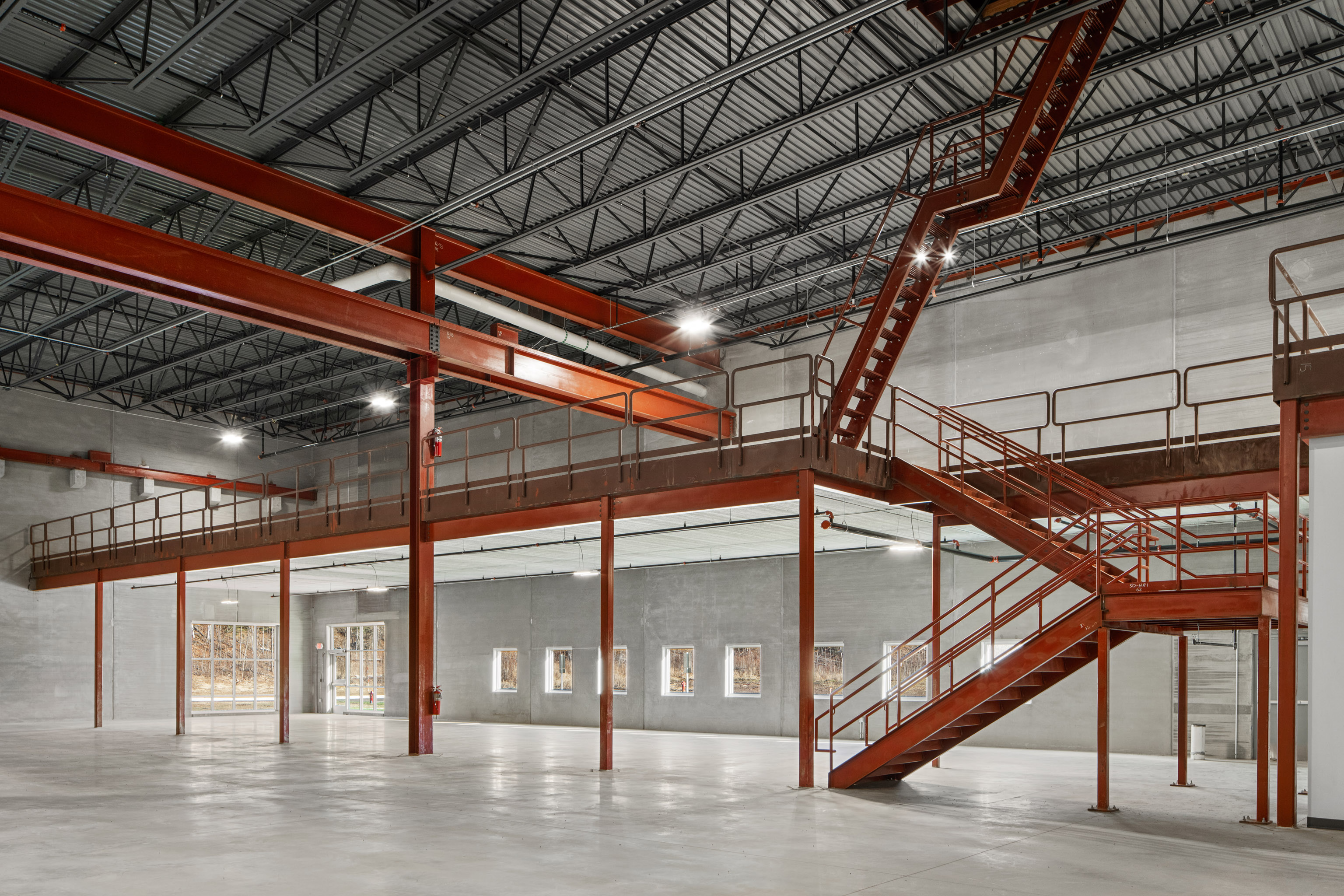 Interior of an empty industrial warehouse with exposed steel beams, a red metal staircase and mezzanine, concrete floors, gray concrete walls, and large windows letting in natural light.