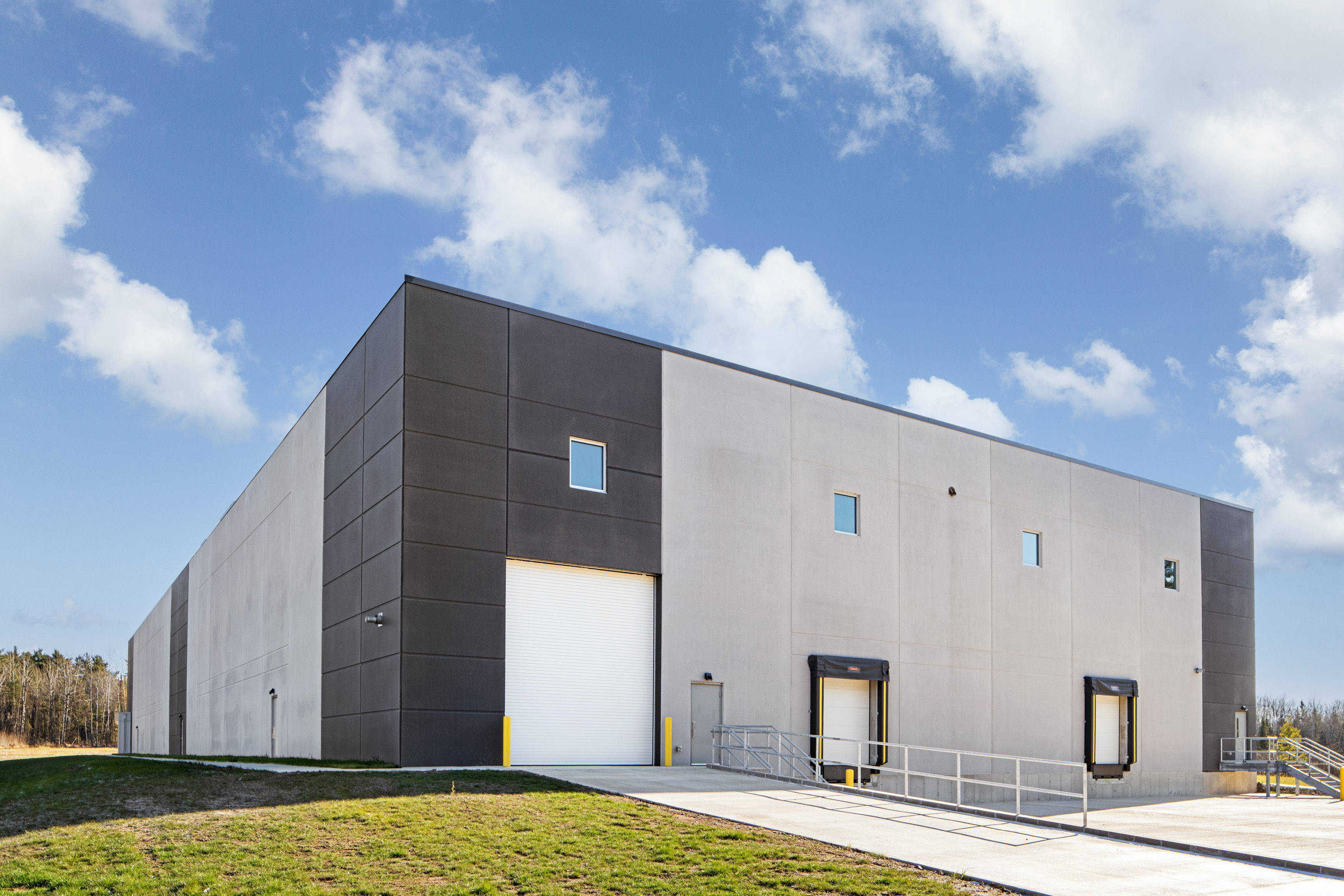 A large, modern warehouse with a white and gray exterior, loading docks, and a roll-up door, surrounded by grass and under a partly cloudy sky.