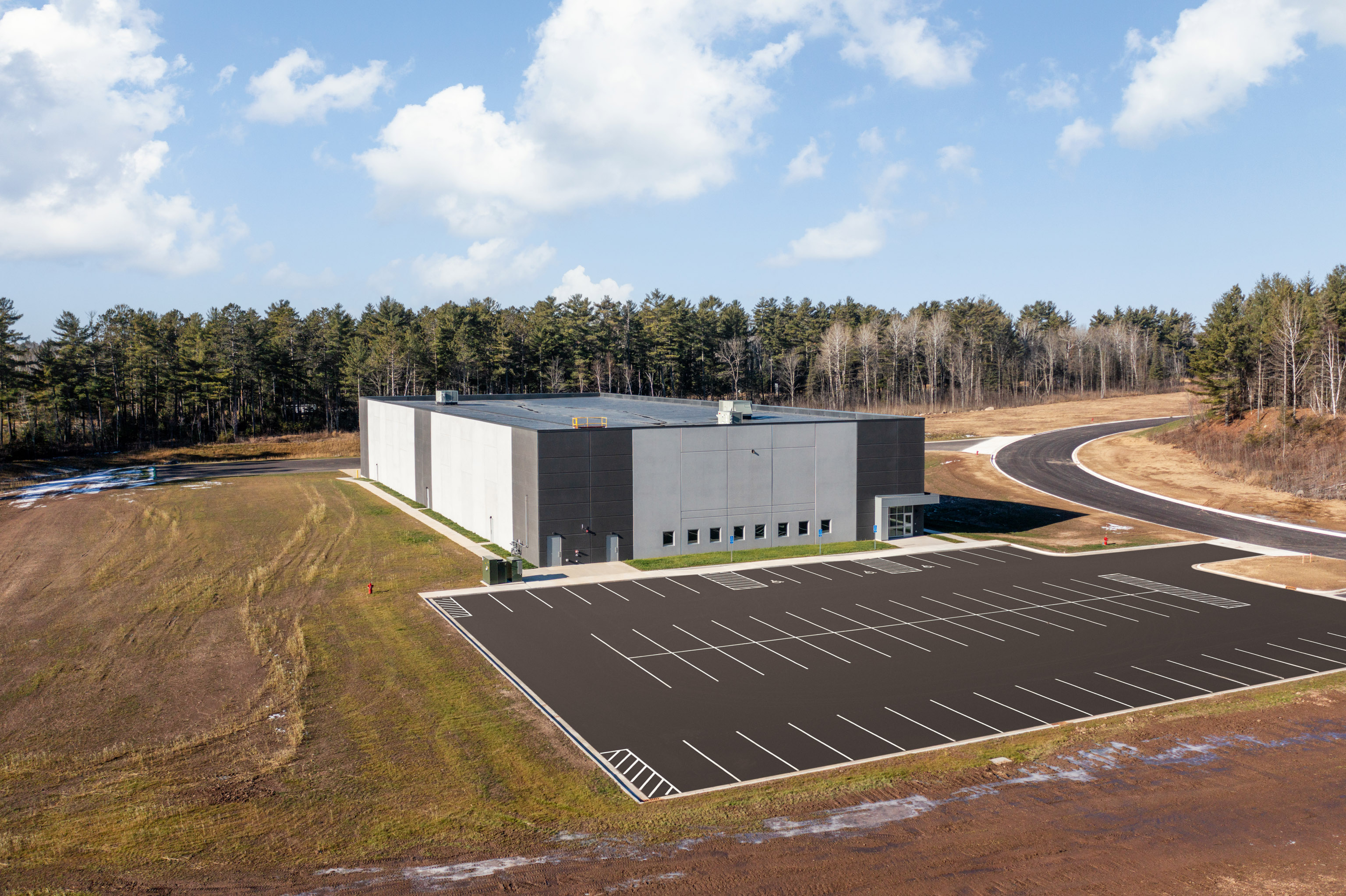 A large, modern warehouse building with a gray exterior sits on a spacious lot, surrounded by trees and an empty parking lot under a partly cloudy sky. A paved road curves near the property.