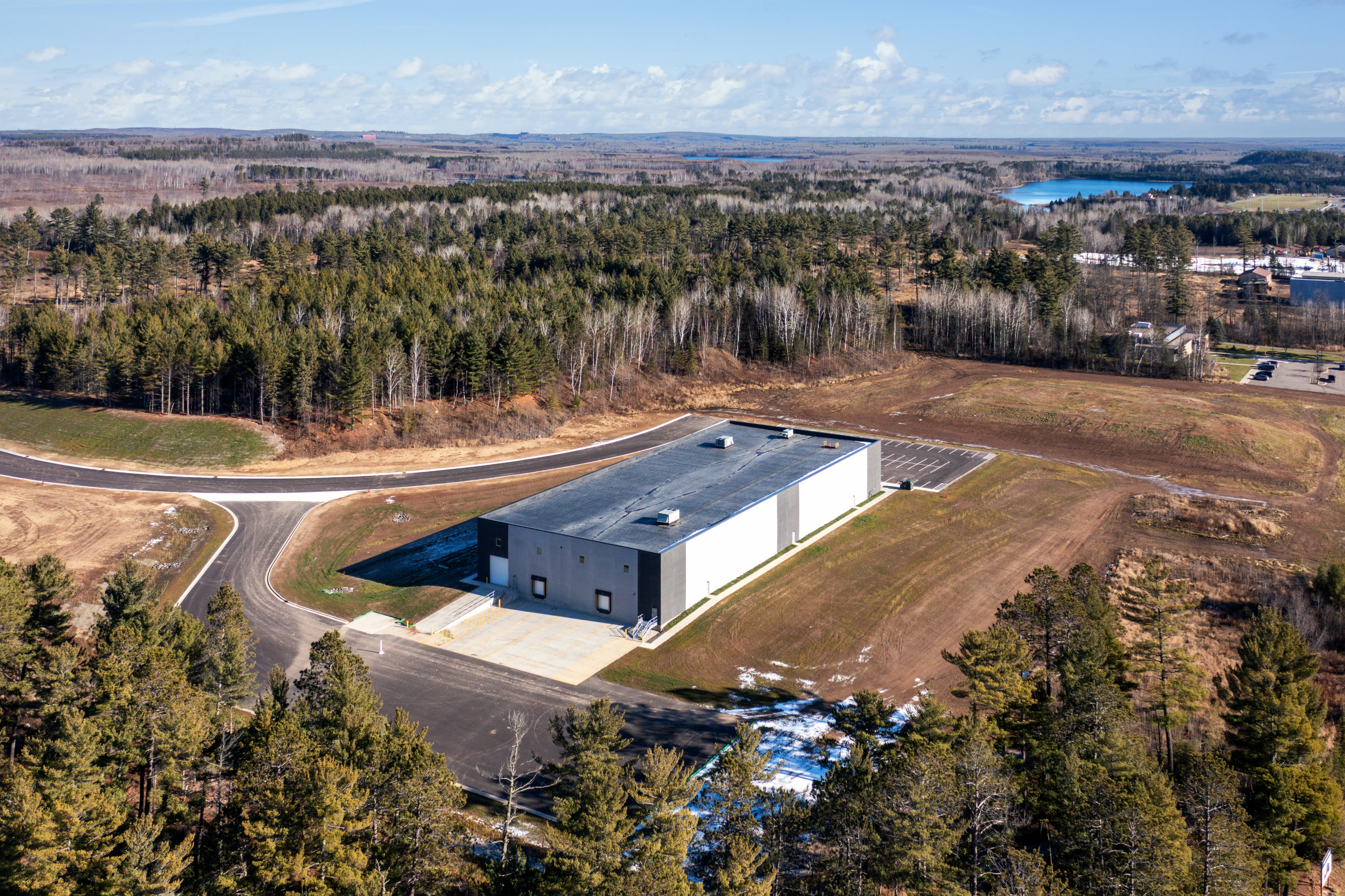 Aerial view of a large, rectangular industrial building surrounded by trees and open fields, with a road curving around the site and a lake visible in the distance.