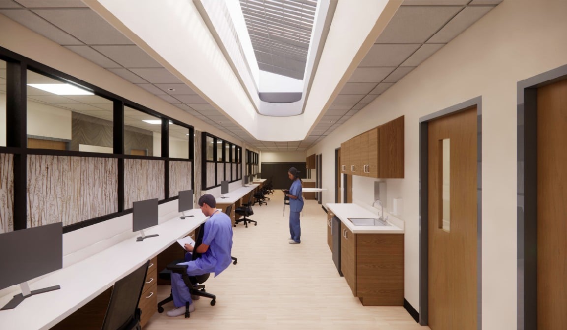 A modern hospital hallway with two medical professionals in scrubs; one is seated at a computer workstation, and the other is standing further down the hall. The area is well-lit with natural light from a skylight above.