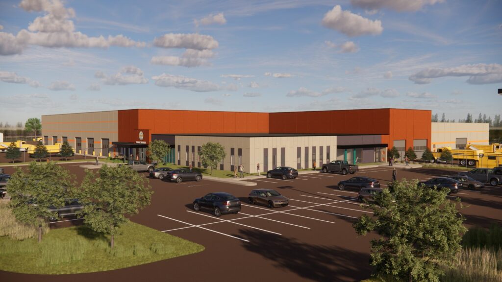 A large, modern industrial building with red and beige exterior walls, a parking lot with cars, trees, and several yellow utility trucks nearby under a partly cloudy sky.