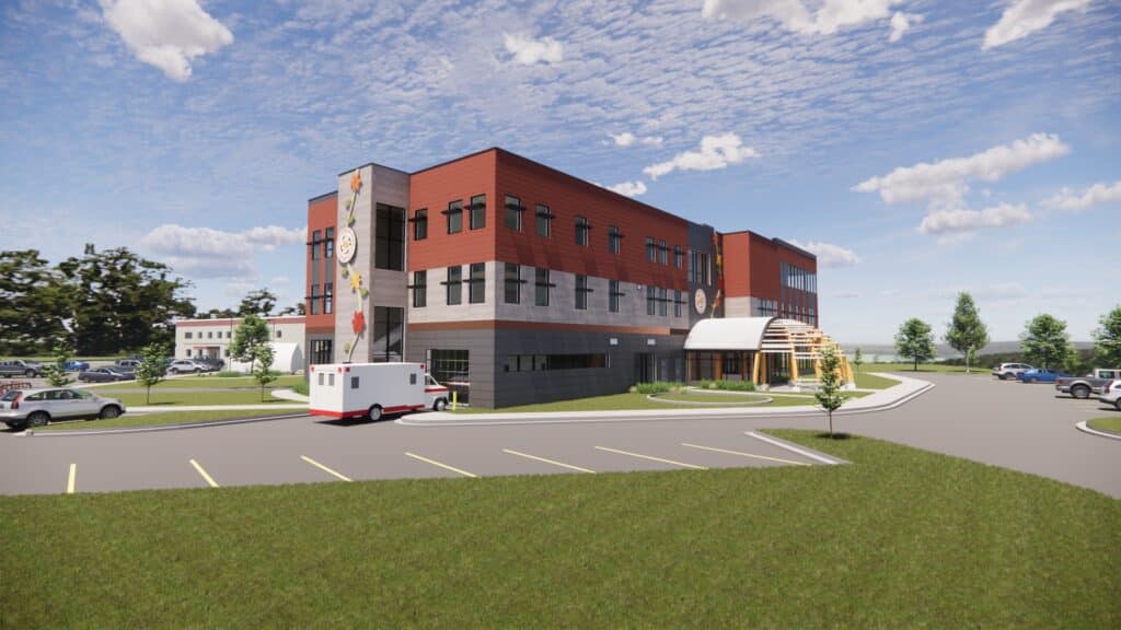 A modern, three-story medical building with red and gray exterior panels welcomes Bay Mills Community College Nursing EMT students; an ambulance is parked outside, near a curved entrance canopy, parking lot, green lawns, and trees under a partly cloudy sky.