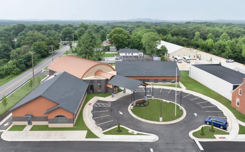 Aerial view of a modern building complex with a circular driveway, parking spaces, and landscaped greenery, surrounded by trees and roads.