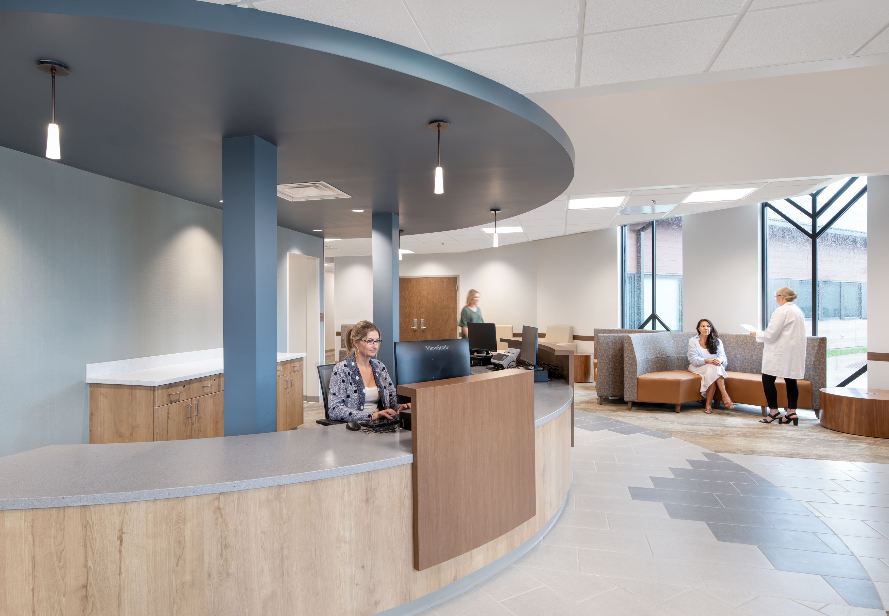 A modern medical office lobby with a curved reception desk, staff at computers, and patients seated in a waiting area near large windows.