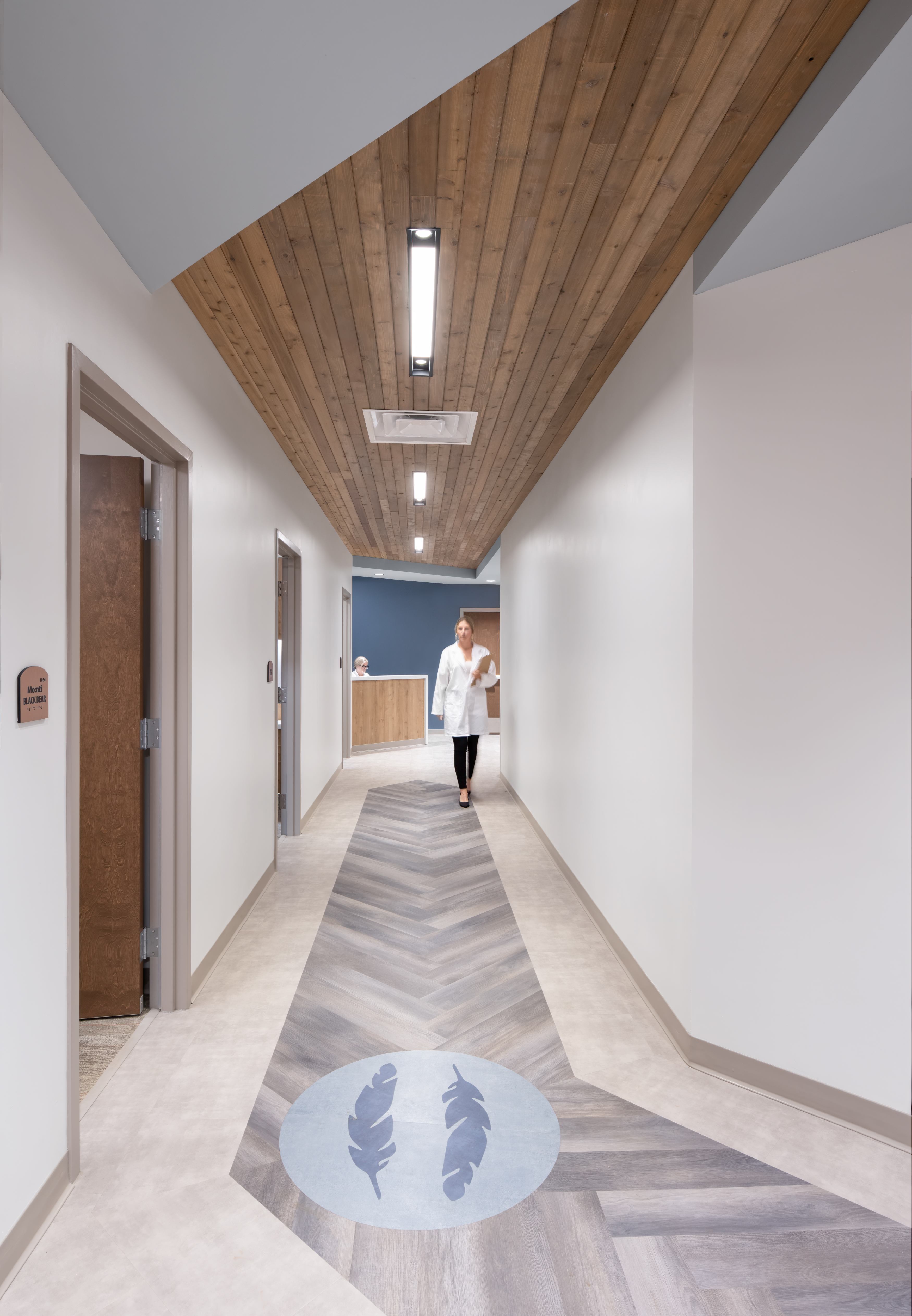 A person in a white coat walks down a modern hallway with a wooden ceiling and feather design on the floor, approaching a reception desk in the background.