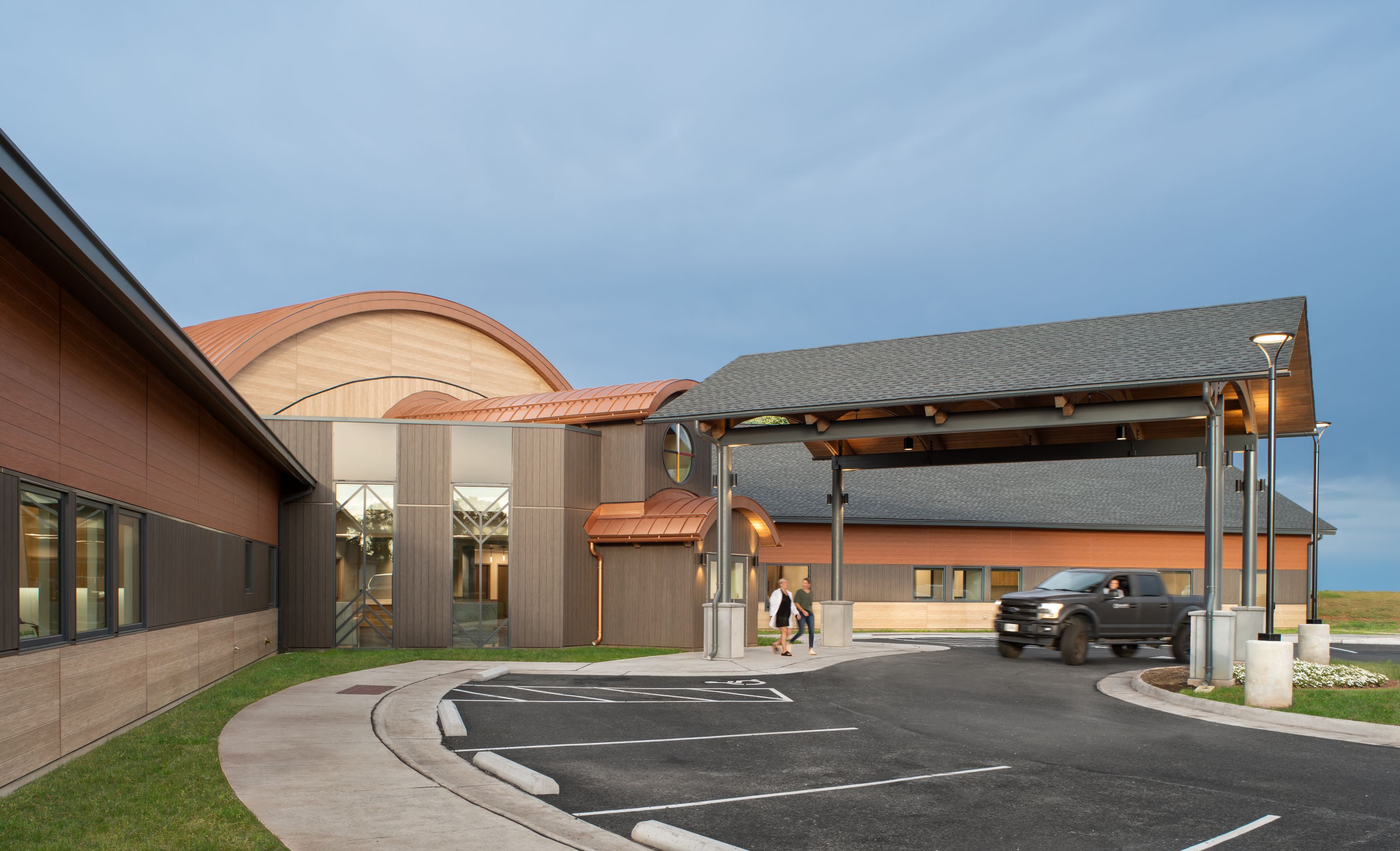 Modern building with wood and metal accents, drive-through entry canopy, parked pickup truck, and two people walking toward the entrance under a cloudy sky.