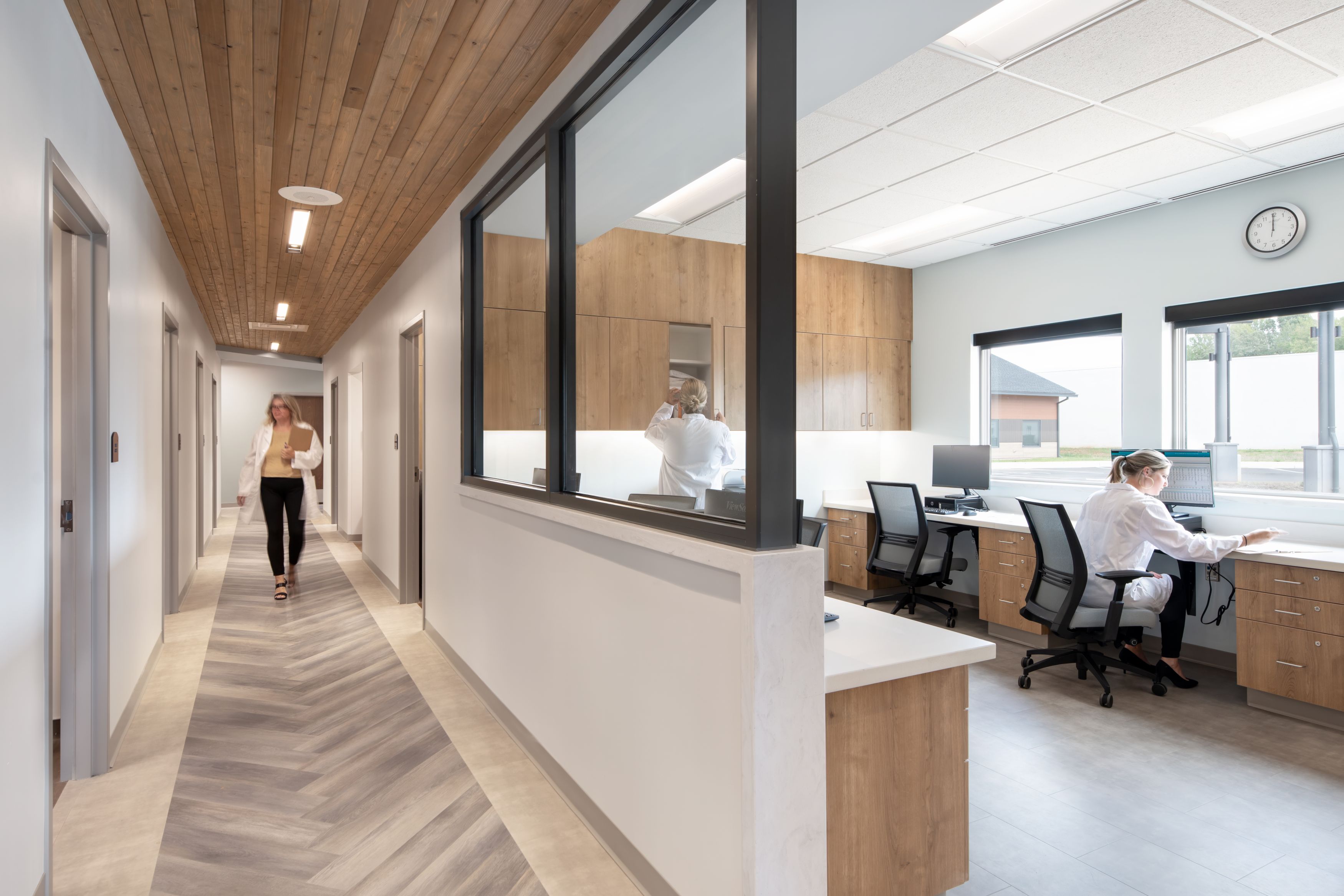 A woman walks down a hallway with wood ceiling and gray chevron floor; on the right, two people in lab coats work at desks in a glass-walled office with computers and wooden cabinets.